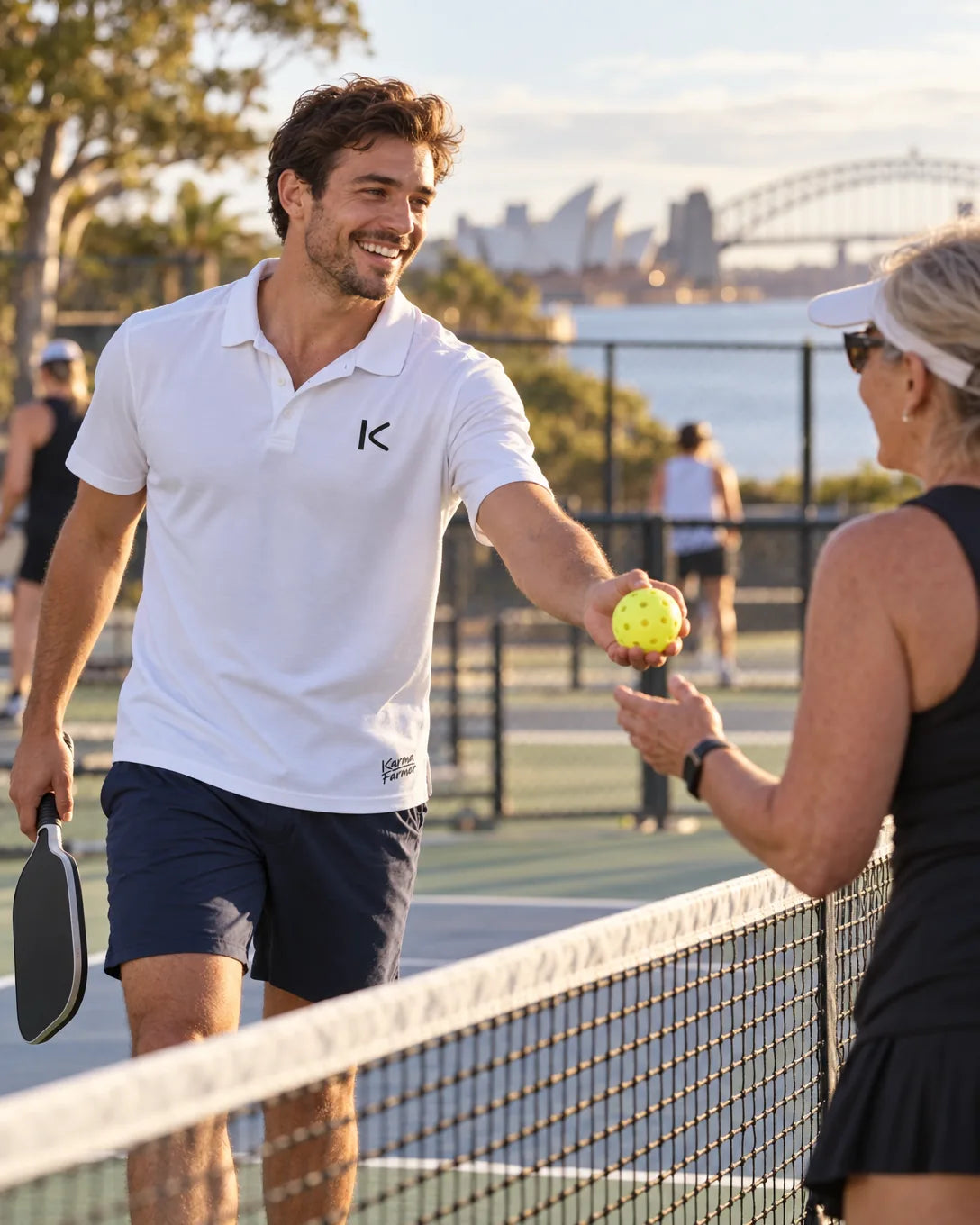 A man in the white Court Polo hands a stray ball back to an older player on the next pickleball court.