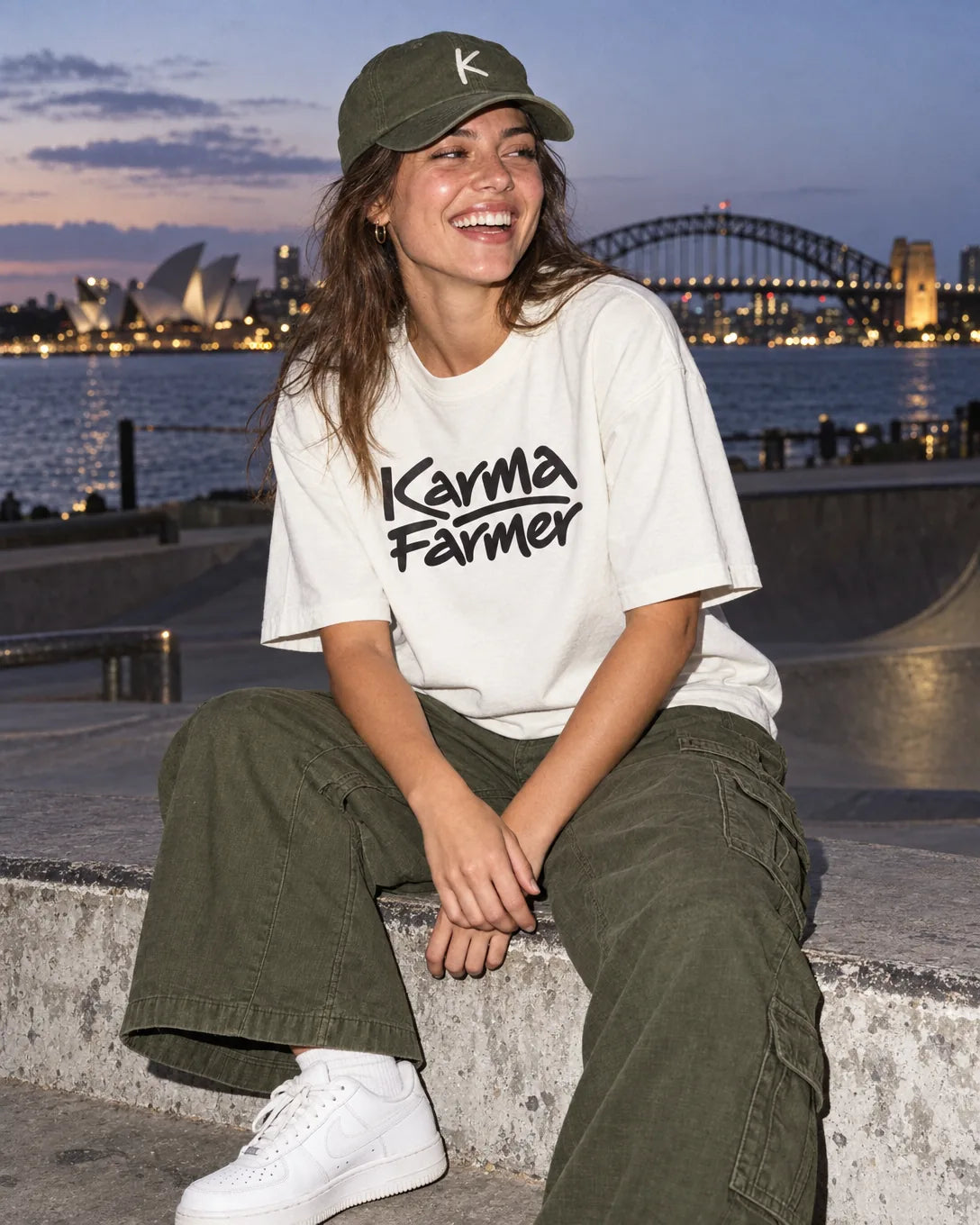 A young woman laughs on a concrete ledge by Sydney Harbour at dusk, in a bone Karma Farmer tee, olive cargo pants and an olive K cap.