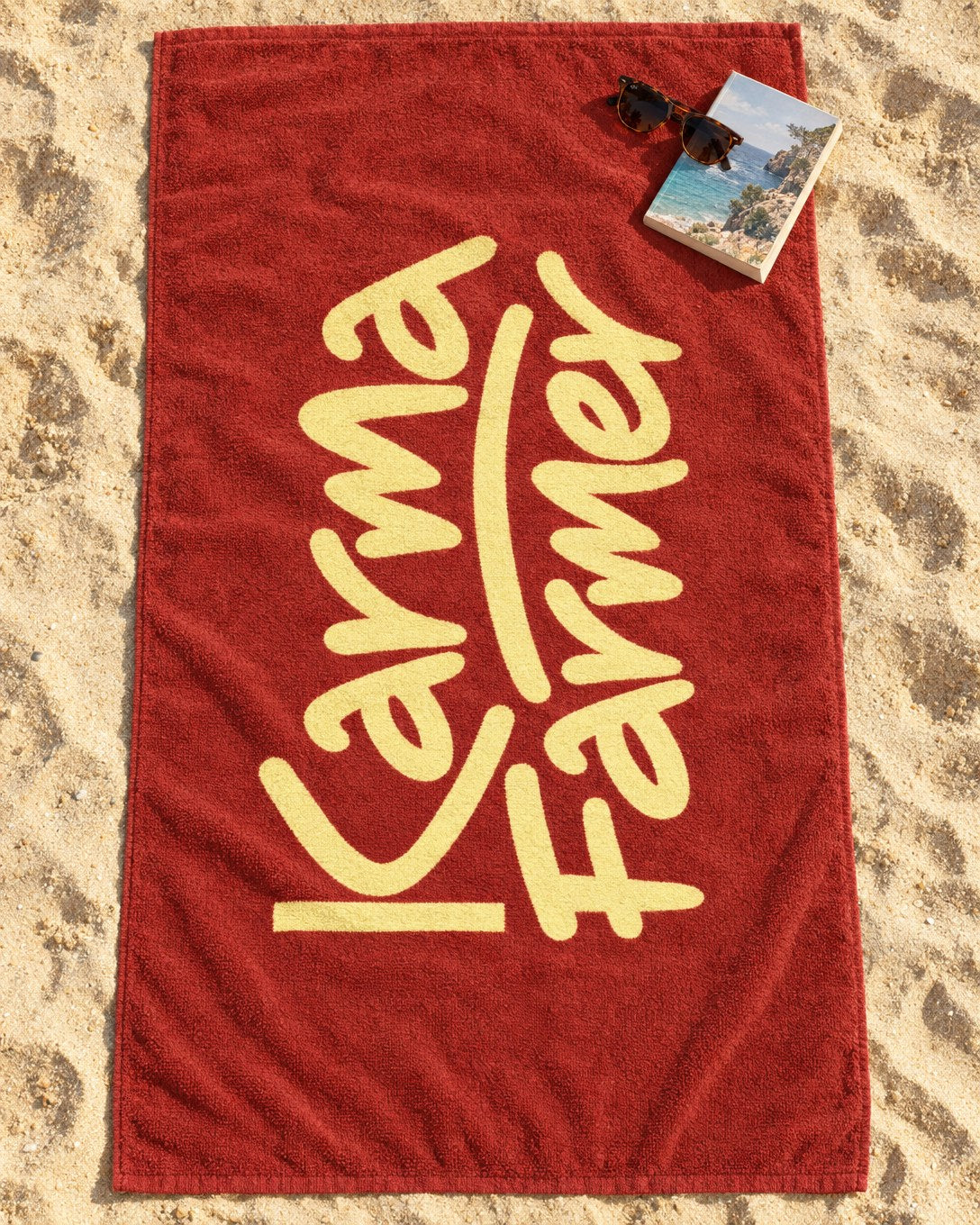 A red Beach Towel on the sand with the Karma Farmer signature in pale yellow along it.
