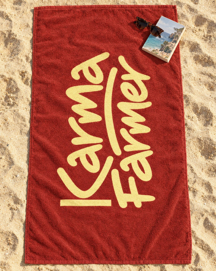 A red Beach Towel on the sand with the Karma Farmer signature in pale yellow along it.