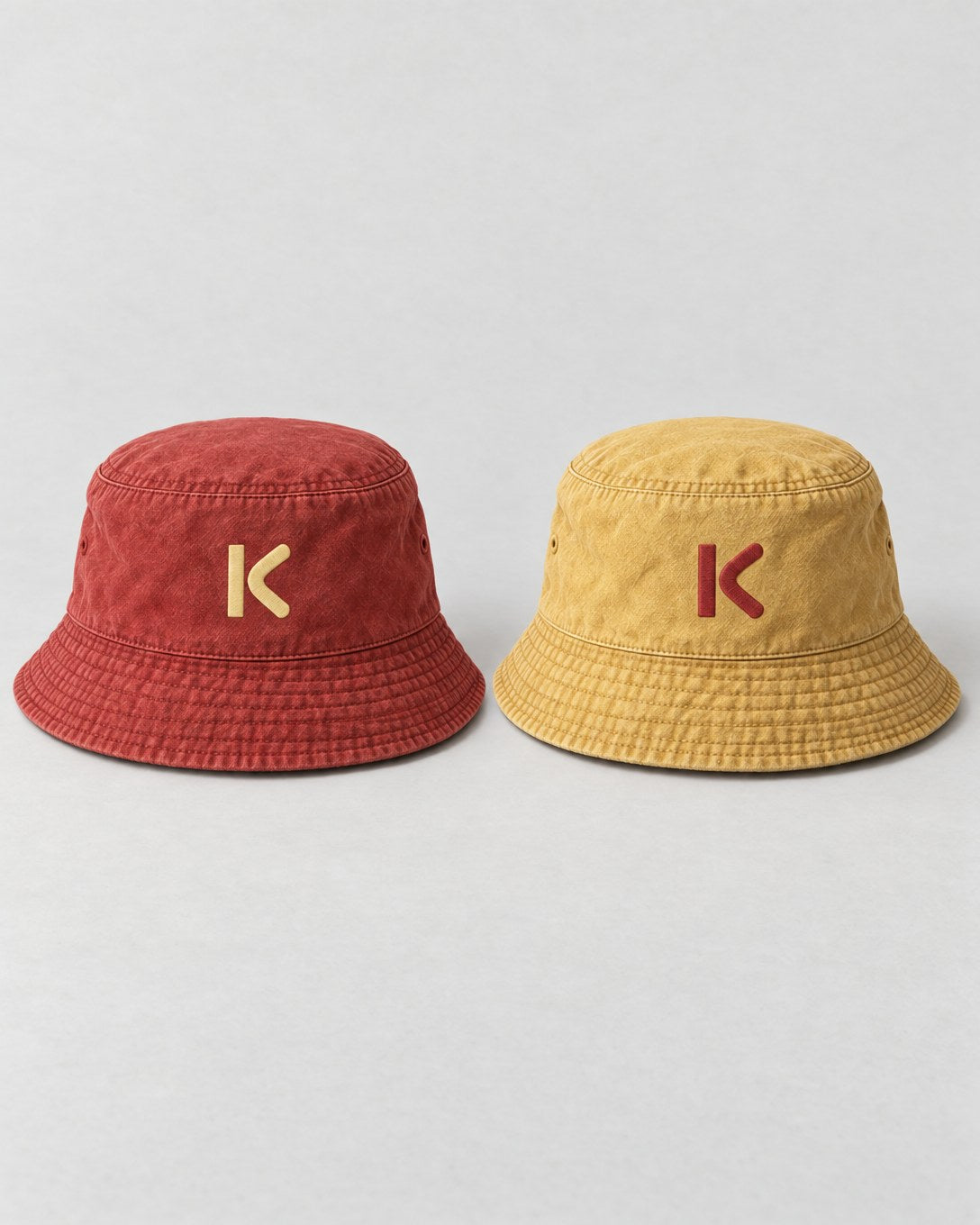 The reversible Bucket Hat shown both ways: brick red with a pale yellow K, and faded mustard with a red K.
