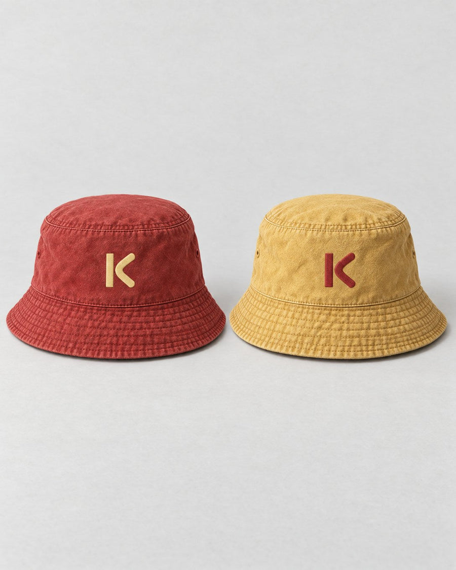 The reversible Bucket Hat shown both ways: brick red with a pale yellow K, and faded mustard with a red K.