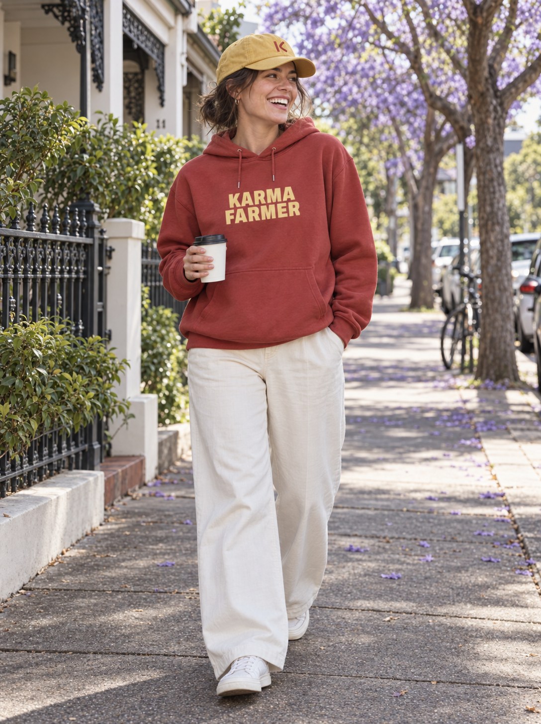 A woman walks down a Sydney street under jacaranda trees in the red Summer Hoodie and a yellow K cap, holding a coffee.