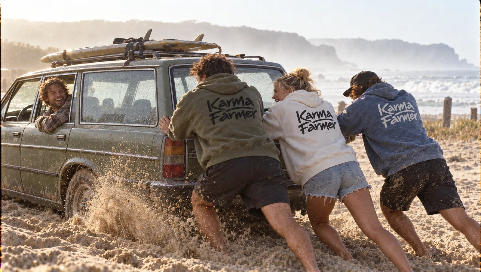 Three friends in Karma Farmer hoodies push a stranger's car out of beach sand at sunrise.