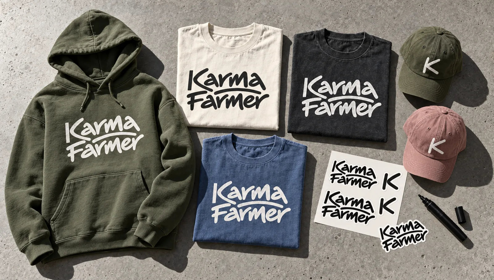 Flat-lay on concrete of an olive hoodie, bone, washed black and washed blue tees printed with the Karma Farmer signature, two K caps, a sticker sheet and a marker.