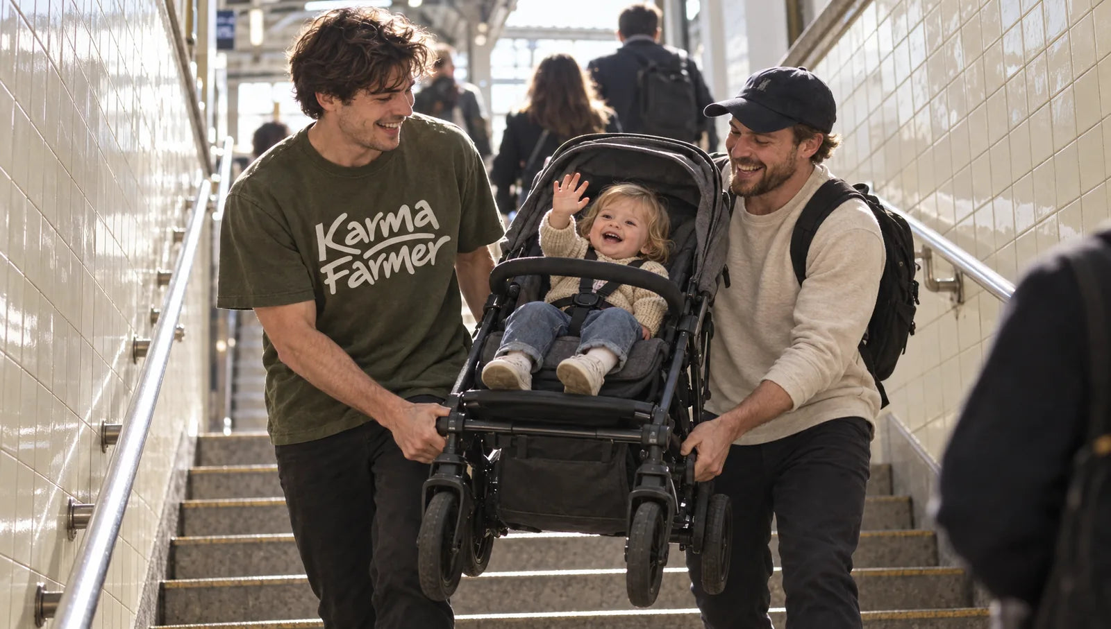 A man in an olive Karma Farmer tee helps a father carry a pram up station stairs.